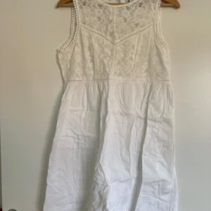 Vit ärmlös spetsklänning - Superfin vit klänning med spets upptill och knappar i ryggen. Ärmlös modell med rund hals och lätt utställd kjol. Perfekt för sommarens alla tillfällen, med romantiska spetsdetaljer och luftig känsla.