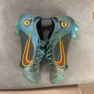 Nike Mercurial fotbollsskor i turkos och orange - Säljer ett par Nike Mercurial fotbollsskor i turkos med orange och gula detaljer. Skorna har snörning och mönstrad ovandel, samt rund tå och platt sula med dobbar för bra grepp på planen. Säljer för att det är för små för mig. Bra skick 