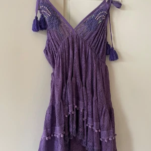 Lila spetsklänning från Antica Sartoria by Giacomo Cinque - Superfin lila klänning i spets med volanger och dekorativa pärlor vid axelbanden. Klänningen har knytband med tofsar på axlarna och v-ringad hals. Perfekt för dig som vill sticka ut med en färgglad och detaljerad look. Köpt i Italien och aldrig använd med prislapp kvar 