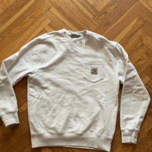 Vit sweatshirt från Carhartt - Säljer en vit sweatshirt från Carhartt med rund halsringning och en bröstficka med logotyp. Tröjan har långa ärmar och ribbade muddar vid ärmslut och nederkant. Perfekt för en avslappnad stil.