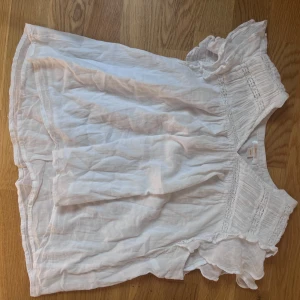 Vit blus med volangdetaljer - Säljer en luftig vit blus med volangdetaljer vid ärmar och axlar. Blusen har korta ärmar och en lös passform, perfekt för varma dagar. Smockad upptill för extra detalj.