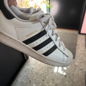 Adidas Superstar vita sneakers med svarta ränder - Klassiska Adidas Superstar sneakers i vitt skinn med svarta ränder och snörning. Skorna har den ikoniska snäckformade tån och Adidas-logga på hälen. Perfekta för en sportig och stilren look.