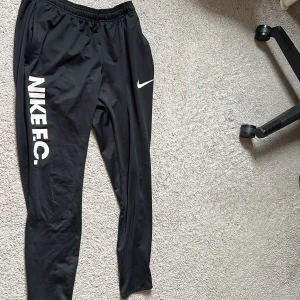 Svarta Nike FC Dri-FIT träningsbyxor - Säljer ett par svarta Nike FC träningsbyxor med Dri-FIT-teknologi. Byxorna har resår i midjan, smal passform och vit Nike-logga samt texten NIKE F.C. längs ena benet. Perfekta för fotboll eller träning.