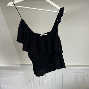 Svart oneshoulder topp från Pull&Bear - Säljer en svart oneshoulder topp från Pull&Bear i storlek XS. Toppen har volangdetalj och är tillverkad i ett lätt, syntetiskt material. Perfekt för varma dagar eller en snygg layering-look.
