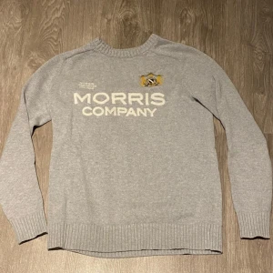 Morris tröja - Säljer en ljusgrå stickad tröja från Morris Company med broderad logga och text framtill. Tröjan har rund halsringning, långa ärmar och ribbade muddar vid ärmslut och nederkant. Perfekt för en stilren och klassisk look.