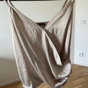 Beige omlottklänning med smala axelband - Säljer en snygg beige omlotttopp med smala axelband och djup v-ringning. Toppen har en draperad framsida och är tillverkad i ett mjukt material som faller fint. Perfekt för varma dagar eller sommarens tillställningar.