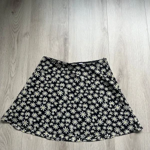 Svart blommig kjol från H&M Divided - Supersöt svart kjol med vita och gula prästkragar från H&M Divided. Kjolen har en lätt utställd form.Bekväm resår i midjan och mjukt bomullsmaterial. Säljer då den inte används längre,är i mycket bra skick. Storlek M 