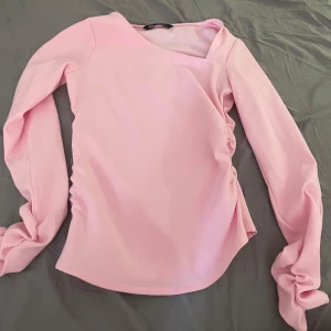 Ljusrosa långärmad topp från Zara - Säljer en ljusrosa långärmad topp från Zara med asymmetrisk halsringning och rynkade detaljer på sidan. Toppen har en modern och stilren design, perfekt för att lyfta din outfit med en pop av färg. Har gått lite sönder men lätt att sy ihop är bild på det 