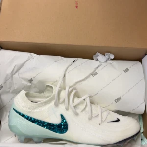 Nike Phantom GX II fotbollsskor - Säljer ett par vita Nike fotbollsskor med en glansig turkos Swoosh på sidan. Skorna har snörning och är designade för att ge bra grepp på planen. Perfekta för dig som vill sticka ut med stil på matchen.