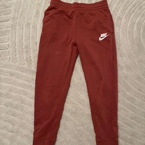 Rosa mjukisbyxor från Nike - Säljer ett par rosa mjukisbyxor från Nike med vit logga på vänster lår. Byxorna har resår i midjan och muddar vid bensluten. Perfekta för en avslappnad stil och riktigt sköna att ha på sig. Kolla min profil för fler kläder!