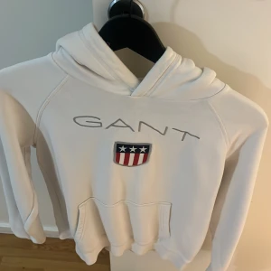 Vit hoodie från GANT - Vit hoodie från GANT med broderad logga och amerikansk flagga på bröstet. Tröjan har huva och en stor magficka framtill. Perfekt för en avslappnad och sportig stil.