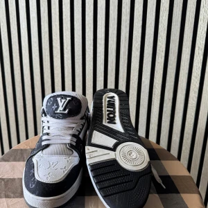 Svarta och vita Louis Vuitton sneakers - Säljer ett par svarta och vita Louis Vuitton sneakers med tydliga LV-loggor och monogramdetaljer. Skorna har snörning, rund tå och platt sula. Materialmix av skinn och syntet, med kontrasterande vita och svarta partier samt präglade mönster på ovandelen.