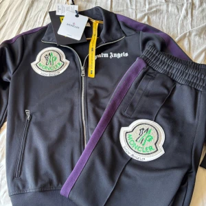 Svart track suit från Moncler x Palm Angels - Säljer en svart track suit från Moncler Genius och Palm Angels med lila ränder på ärmarna, broderade patches på bröstet och armen samt texten 'Mind Control' på ryggen. Jackan har dragkedja och ståkrage. Byxorna har samma lila ränder och samma patches.