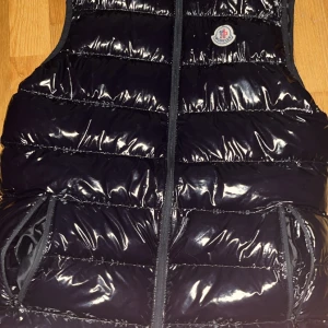 Svart dunväst från Moncler - Säljer en glansig svart dunväst från Moncler med dragkedja framtill och två fickor. Västen har Moncler-logga på bröstet och är quiltad för extra värme. Perfekt för lager-på-lager under kyliga dagar dragkedjan är lite sönder tar 100 kr extra om man vill att jag ska fixa den 