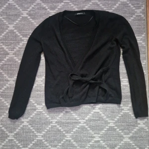 Svart omlottkofta med knyt - Svart omlottkofta från Gina Tricot med knyt i midjan. Långärmad och i mjukt, tunt stickat material. Perfekt att slänga över en topp för en enkel och stilren look.