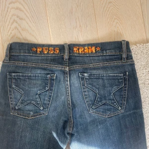 Blå jeans med stjärnfickor och broderi - Unika blå jeans med stjärnformade bakfickor och broderad text 'PUSS' och 'KRAM' i orange på linningen. Klassisk femficksmodell med normal passform och bootcut-ben. Perfekta för dig som vill sticka ut med coola detaljer. midjemått: 38cm