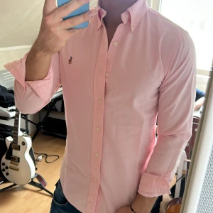 Ljusrosa skjorta från Ralph Lauren - Säljer en ljusrosa skjorta från Ralph Lauren med klassisk button-down krage och broderad logga på bröstet. Skjortan har långa ärmar och knäppning framtill. Perfekt för en stilren och fräsch look.