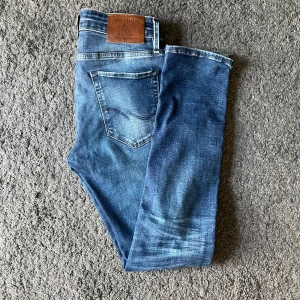 Blå slitna jeansbyxor  - Säljer ett par blå jeansbyxor från jack and jones med slitna detaljer och klassisk femficksdesign. Jeansen har normal passform och raka ben, samt läderpatch med logga bak i midjan. Perfekta för en avslappnad och trendig stil.