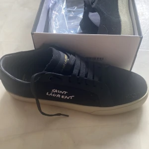  Saint Laurent storlek 42 - Säljer ett par svarta sneakers från Saint Laurent med vit sula och svart snörning. Skorna har en stilren design med Saint Laurent-logga broderad i vitt på sidan. Perfekta för dig som gillar klassiska och trendiga sneakers. Storlek 42 är skorna och har ni några funderingar hör av er.