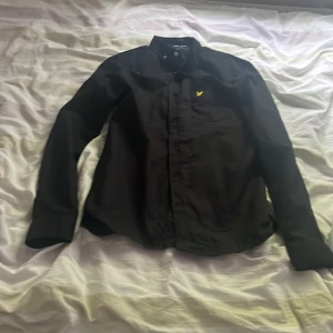 Svart skjorta från Lyle & Scott - Säljer en svart skjorta från Lyle & Scott med klassisk krage och knappstängning framtill. På bröstet finns den ikoniska broderade gula örnen. Perfekt för dig som gillar stilrena plagg med diskreta detaljer.