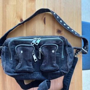 Svart axelväska i manchester med dragkedjor - Cool svart axelväska i manchestertyg med flera silvriga dragkedjor och nitar på axelbandet. Väskan har två mindre fickor framtill och justerbart axelband. Perfekt för att bära dina viktigaste saker med stil.