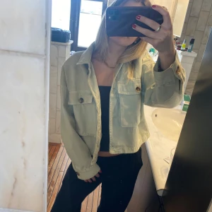 Grön croppad jeansjacka - Säljer en grön croppad jeansjacka med stora bröstfickor och knappar framtill. Jackan har en avslappnad passform och långa ärmar. Perfekt att slänga över en topp för en cool look.