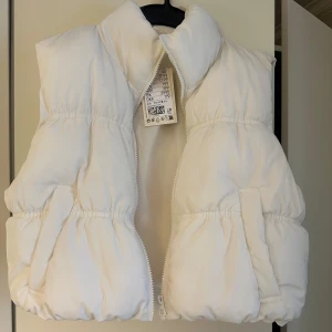 Vit dunväst  - Säljer en fluffig och trendig vit dunväst från Divided. Västen har hög krage, dragkedja framtill och två fickor på sidorna.Perfekt att styla över en hoodie eller långärmad tröja. Storlek S (pris går att diskuteras) 