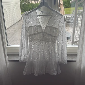 Vit broderad kortklänning med volang - Superfin vit kortklänning med broderade detaljer och volang nertill. Klänningen har långärmade ärmar, v-ringning och knyt framtill. Perfekt för sommarens alla tillfällen! Har bara används en gång och är i toppenskick. 