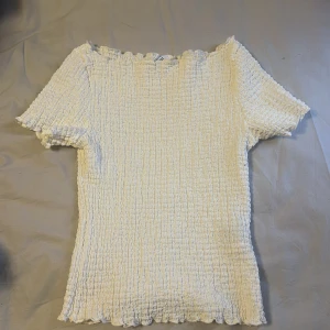 Vit smockad topp - Säljer en vit smockad topp med korta ärmar och vågig kant vid halsringning och nederkant. Toppen är stretchig och har en tight passform, perfekt att styla till olika outfits. Materialet är mjukt och bekvämt.