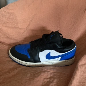 Nike Air Jordan 1 Low blå och svart - Säljer en Nike Air Jordan 1 Low med blå, svart och vit färgkombination. Skon har klassisk snörning, rund tå och platt sula. Ikonisk Nike Swoosh i blått på sidan och svart ovandel i skinn. Perfekt för dig som gillar streetwear och sneakers.
