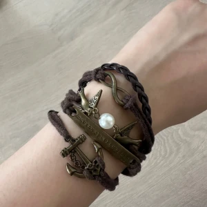 Brunt armband med fåglar och pärldetalj - Unikt armband med flera bruna band, antikguldiga detaljer som fåglar, oändlighetssymbol, ankare och en vit pärla. Armbandet har även en flätad sektion och textplatta. Perfekt accessoar för dig som gillar boho-stil.