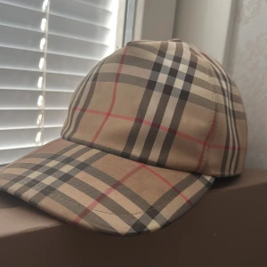 Burberry  - Säljer en klassisk beige keps från Burberry med det ikoniska rutmönstret i svart, rött och vitt. Kepsen har justerbar baksida och en svart patch med texten 'Burberry London England'. Perfekt accessoar för en trendig look.