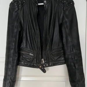 Svart skinnjacka med nitar från Rock'n Blue - Cool svart skinnjacka från Rock'n Blue med silvriga dragkedjor och nitar på axlarna. Jackan har klassisk bikerstil, ståkrage och detaljerade sömmar. Lädret har en tydlig patina vilket jag har försökt fota så gott det går. Perfekt för dig som vill ha en edgy look.