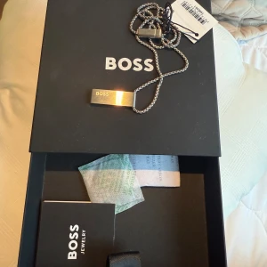 Silverfärgat halsband från BOSS - Stilrent halsband från BOSS i silverfärg med en rektangulär platta där BOSS-loggan är ingraverad. Kedjan är av klassisk modell och halsbandet levereras i originalask med märkning och etikett.