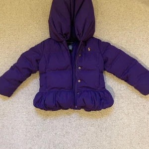 Lila dunjacka med volang från Ralph Lauren - Superfin lila dunjacka från Ralph Lauren med huva och volangdetalj nedtill. Jackan har tryckknappar framtill och är vadderad för extra värme. Perfekt för kyliga dagar och snygg detalj med liten logga på bröstet. Så fin och puffig😊