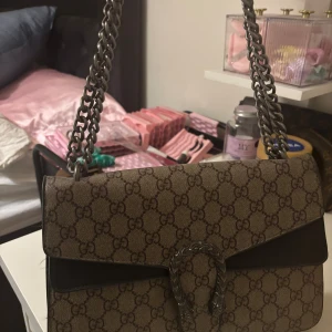  axelväska med kedjerem - Säljer en klassisk axelväska i beige och brunt silverfärgad kedjerem. Väskan har ett dekorativt spänne framtill och detaljer i mörkbrunt skinn. Perfekt för dig som vill ha en tidlös och lyxig accessoar.