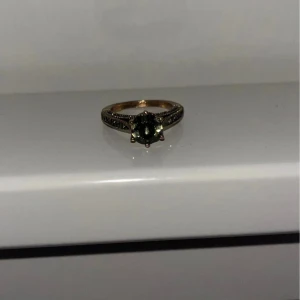 Ring med mörk sten och detaljerad infattning - Snygg ring med en stor mörk sten i mitten och detaljer runt hela bandet. Ringen har en klassisk och elegant design med guldfärgad bas och dekorativa inslag.