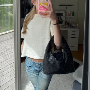 Svart axelväska i flätad syntet - Säljer en svart axelväska med flätad design. Väskan har en mjuk, rundad form och ett brett handtag som gör den bekväm att bära på axeln. Perfekt för att få plats med det viktigaste till vardags.