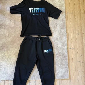 Svart mjukisset från Trapstar - Svarta mjukisbyxor och t shirt från Trapstar med logga i vitt och blått. Byxorna har resår och snörning i midjan samt sidofickor. Dragkedja vid vaderna, dragkedjan på vänster ben är dock sönder 