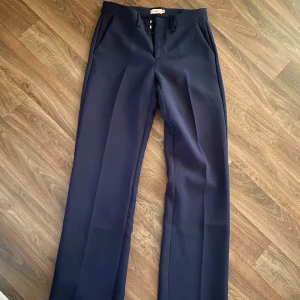 Mörkblå kostymbyxor från Nelly - Säljer ett par mörkblå kostymbyxor från Nelly med klassisk rak passform med en liten utsvängning och pressveck. Byxorna har hällor för bälte och stängs med dragkedja och hyska. Perfekta för en stilren look. Burit de en gång och är i nytt skick. 🩵