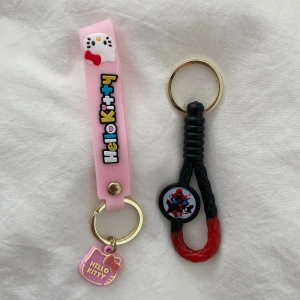 Hello Kitty och Spiderman nyckelringar - Två nyckelringar: en rosa Hello Kitty med text, figur och berlock, samt en svart och röd Spiderman-nyckelring med rund dekoration. Perfekt som accessoar till väskan eller nycklarna.