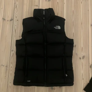 Svart dunväst från The North Face - Säljer en svart dunväst från The North Face med hög krage och dragkedja framtill. Västen har en klassisk puffer-design och logga på bröstet. Perfekt för lager-på-lager och håller dig varm under kyliga dagar.