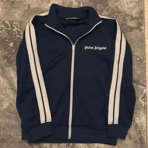 Blå track jacket från Palm Angels - Säljer en mörkblå track jacket från Palm Angels med vita ränder längs ärmarna och vit dragkedja. Jackan har hög krage och logga på bröstet. Perfekt för en sportig och trendig look.