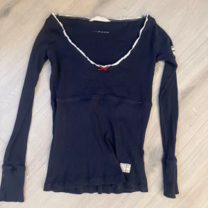 Mörkblå ribbad långärmad topp från Odd Molly - Supersöt mörkblå långärmad topp från Odd Molly med vit spetskant vid ringningen och en liten röd rosett framtill. Tröjan är ribbad och har en feminin v-ringning. Perfekt för dig som gillar detaljer och mjuka material.
