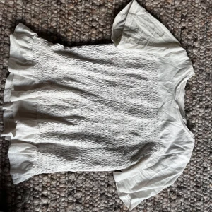Vit blus med smock från Gina Tricot - Säljer en söt vit blus från Gina Tricot med smockad front och volangdetaljer nedtill. Blusen har korta ärmar och rund halsringning. Perfekt för en fräsch och stilren look. 