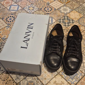 Svarta Lanvin skor - Säljer ett par svarta sneakers från Lanvin med snygg snörning och diskret präglat mönster på tån. Skorna har rund tå och platt sula, tillverkade i skinn och mocka för en lyxig känsla. Perfekta för dig som gillar stilrena och exklusiva sneakers. Använda med försiktighet och fortfarande i bra skick.