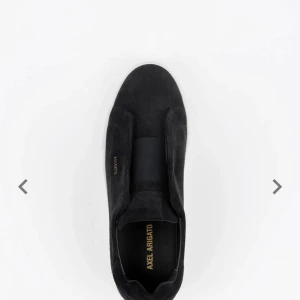 Svarta slip-on sneakers från Axel Arigato - Stilrena svarta slip-on sneakers från Axel Arigato med ovandel i mocka och diskret guldfärgad logga på sidan. Skorna har en vit platt sula och minimalistisk design utan snörning. Kvitto finns har använts 2-3 gånger .