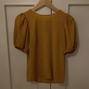 Gul blus med puffärmar - Säljer en snygg gul blus med puffiga korta ärmar och rund halsringning. Blusen har en knappdetalj i nacken och är tillverkad i ett lätt, strukturerat tyg. Perfekt för att liva upp garderoben!