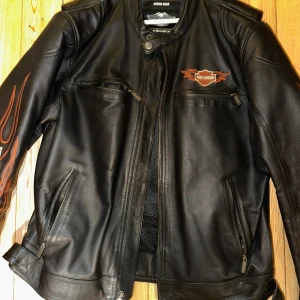 Svart skinnjacka från Harley-Davidson med flames - Säljer en svart skinnjacka från Harley-Davidson med broderade flames i orange och vita loggor på ärmar, bröst och rygg. Jackan har dragkedjor, ståkrage och flera fickor framtill. Perfekt för dig som gillar MC-stil.