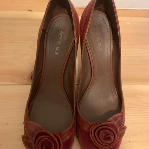 Röda pumps med rosettdetalj från Shoe Biz - Snygga röda pumps i mocka från Shoe Biz med dekorativ rosett framtill. Skorna har klack och rund tå, säger st40 men skulle säga att de även passar 39